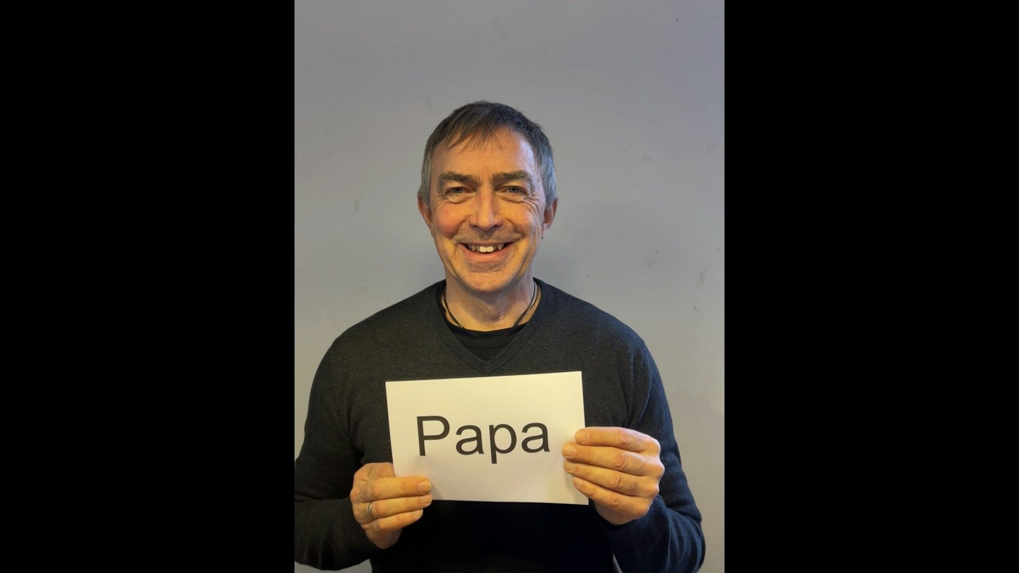 Der Vater hält einen Zettel vor der Brust mit dem Schriftzug „Papa“.