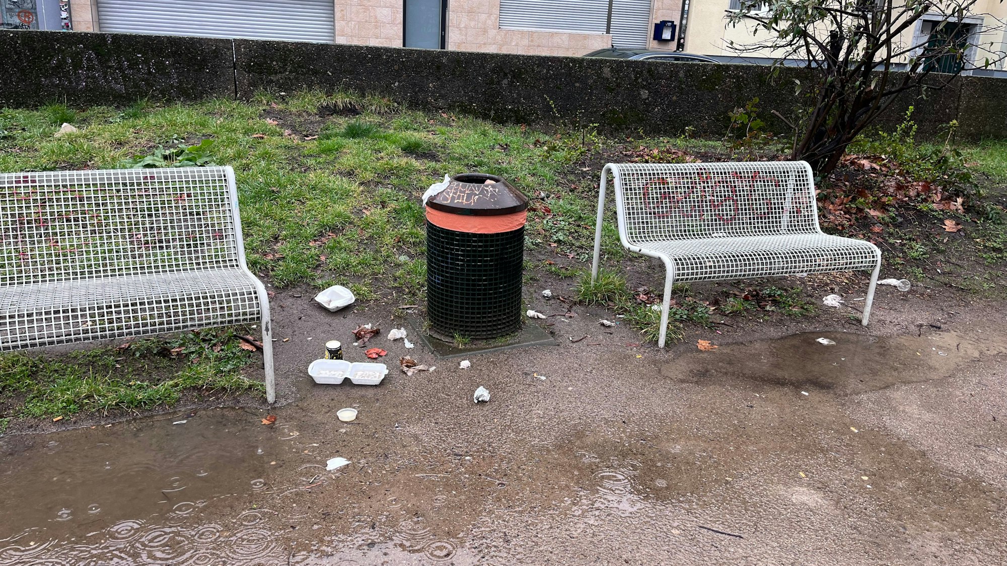 Blick auf zwei Bänke und einen Mülleimer, neben dem Verpackungen liegen.