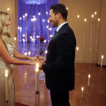 Eine Frau mit blonden Haaren steht vor einem Mann im Anzug, ihre Hände sind in seine gelegt.