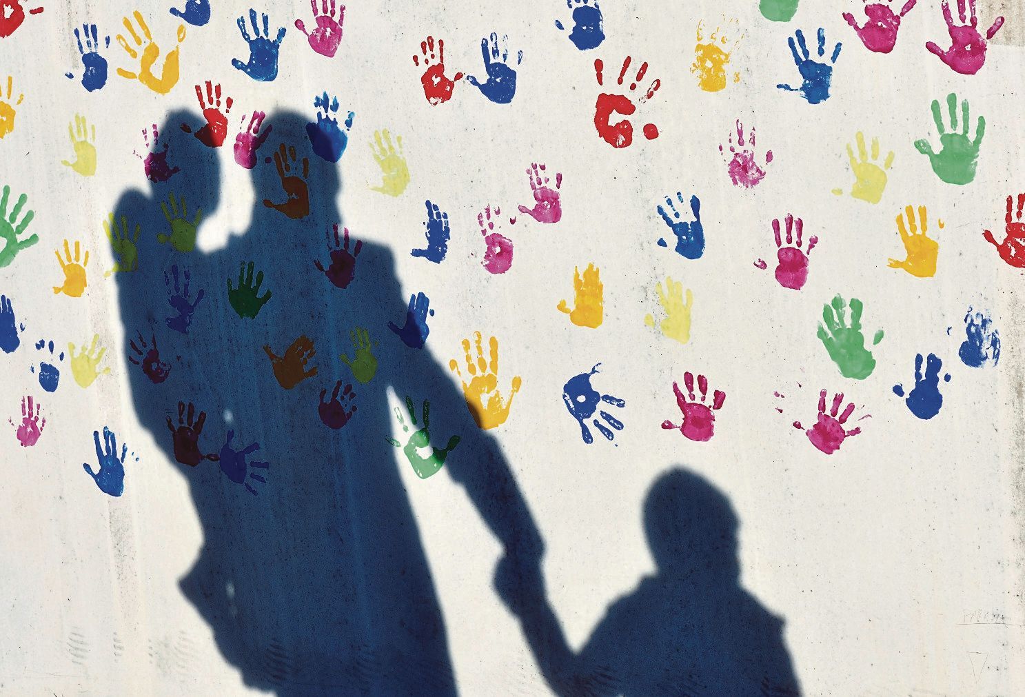 Ein Mann mit einem Kind auf dem Arm und einem an der Hand wirft einen Schatten auf eine mit bunten Handabdrücken bemalte Wand einer Kindertagesstätte.