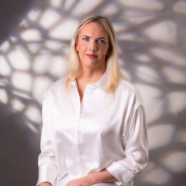 Julia Monro steht vor einer weißen Wand, auf die Schatten fallen. Sie trägt eine weße Bluse und lächelt in die Kamera.
