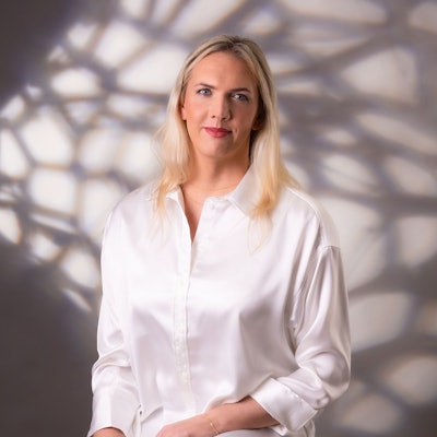 Julia Monro steht vor einer weißen Wand, auf die Schatten fallen. Sie trägt eine weße Bluse und lächelt in die Kamera.