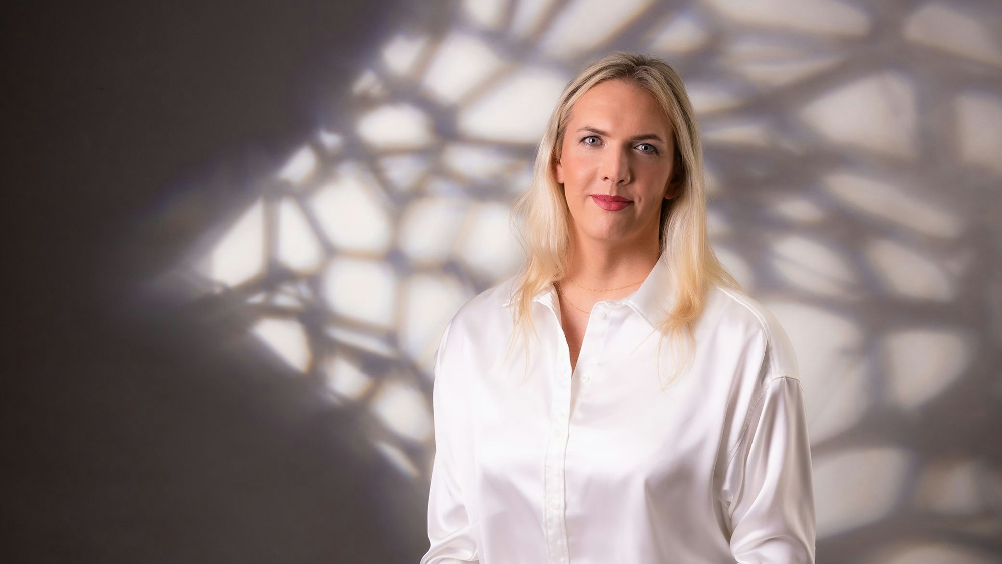 Julia Monro steht vor einer weißen Wand, auf die Schatten fallen. Sie trägt eine weße Bluse und lächelt in die Kamera.