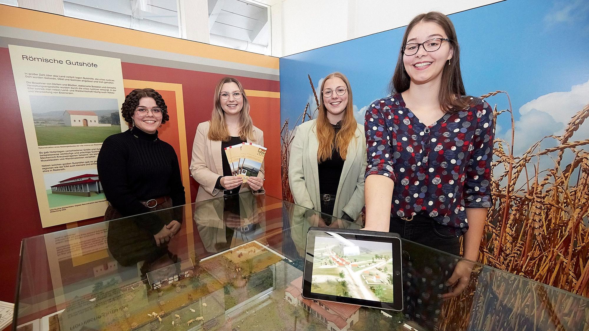 Vier junge Frauen stehen in einem Ausstellungsraum an einem Glastisch, eine hält ein Tablet in der Hand.
