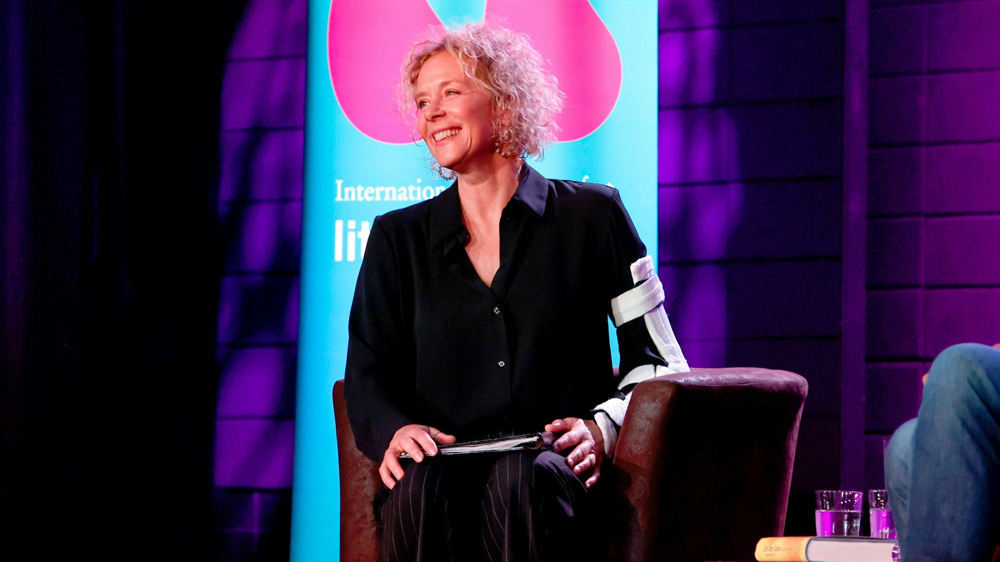 Katja Riemann trägt ein schwarzes Hemd und lächelt. Sie hat eine Schiene am linken Arm. Im Hintergrund ein blaues Banner der lit.Cologne und violette Beleuchtung.