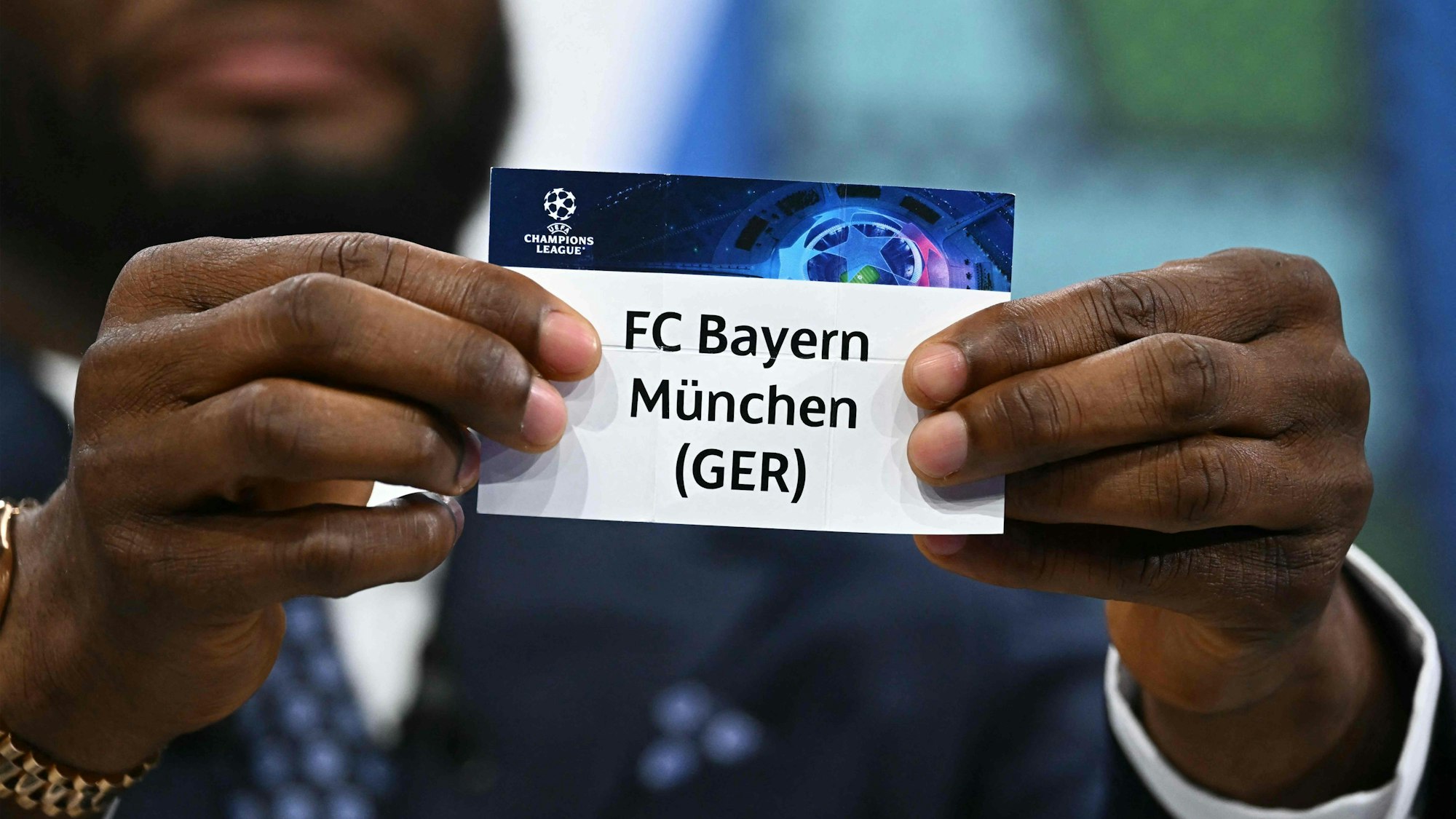 John Obi Mikel hält den Loszettel des FC Bayern München in die Kamera.