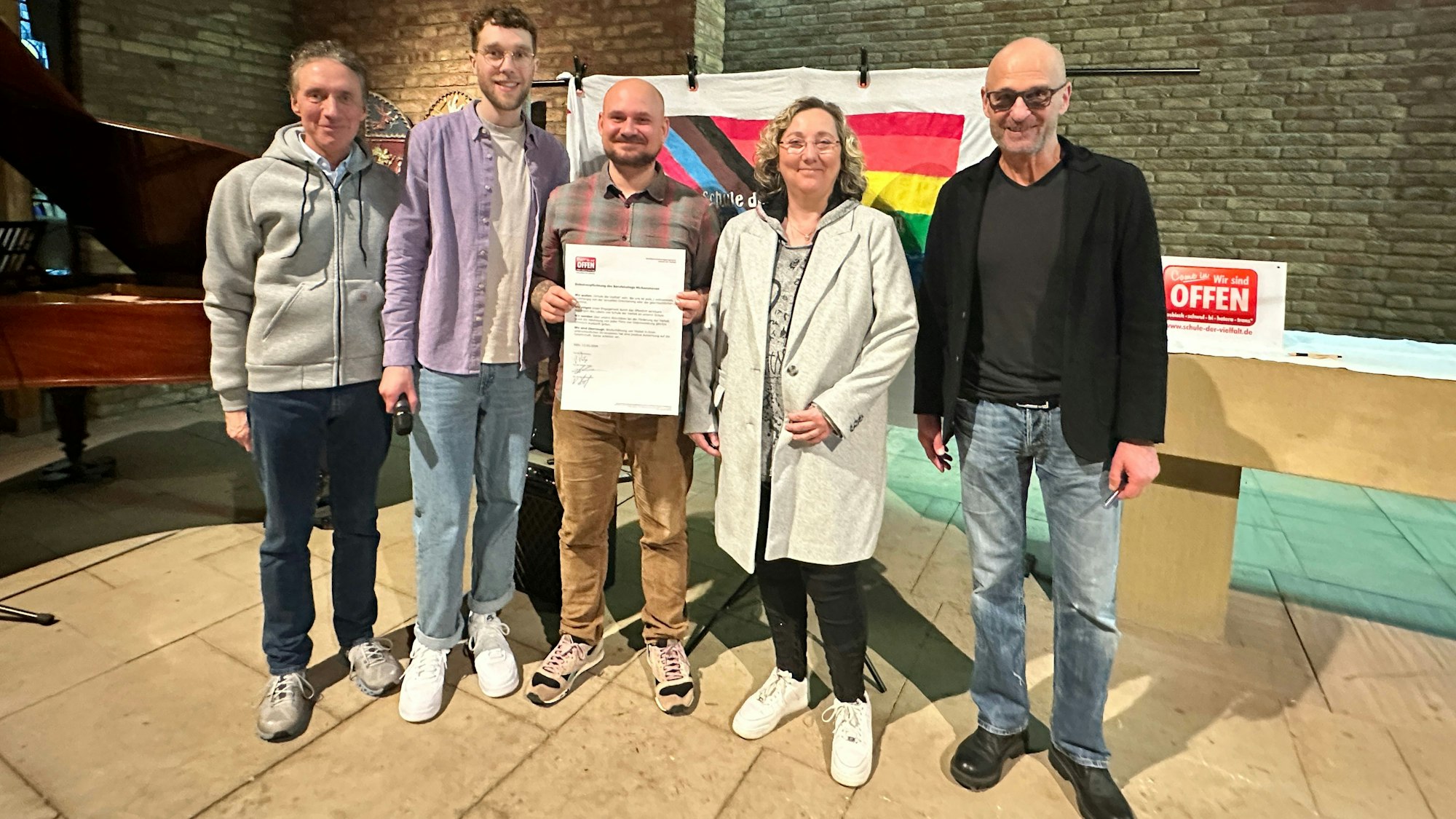 Vier Männer und eine Frau stehen nebeneinander, einer hält einen Vertrag in der Hand, der überdimensioniert groß ist.