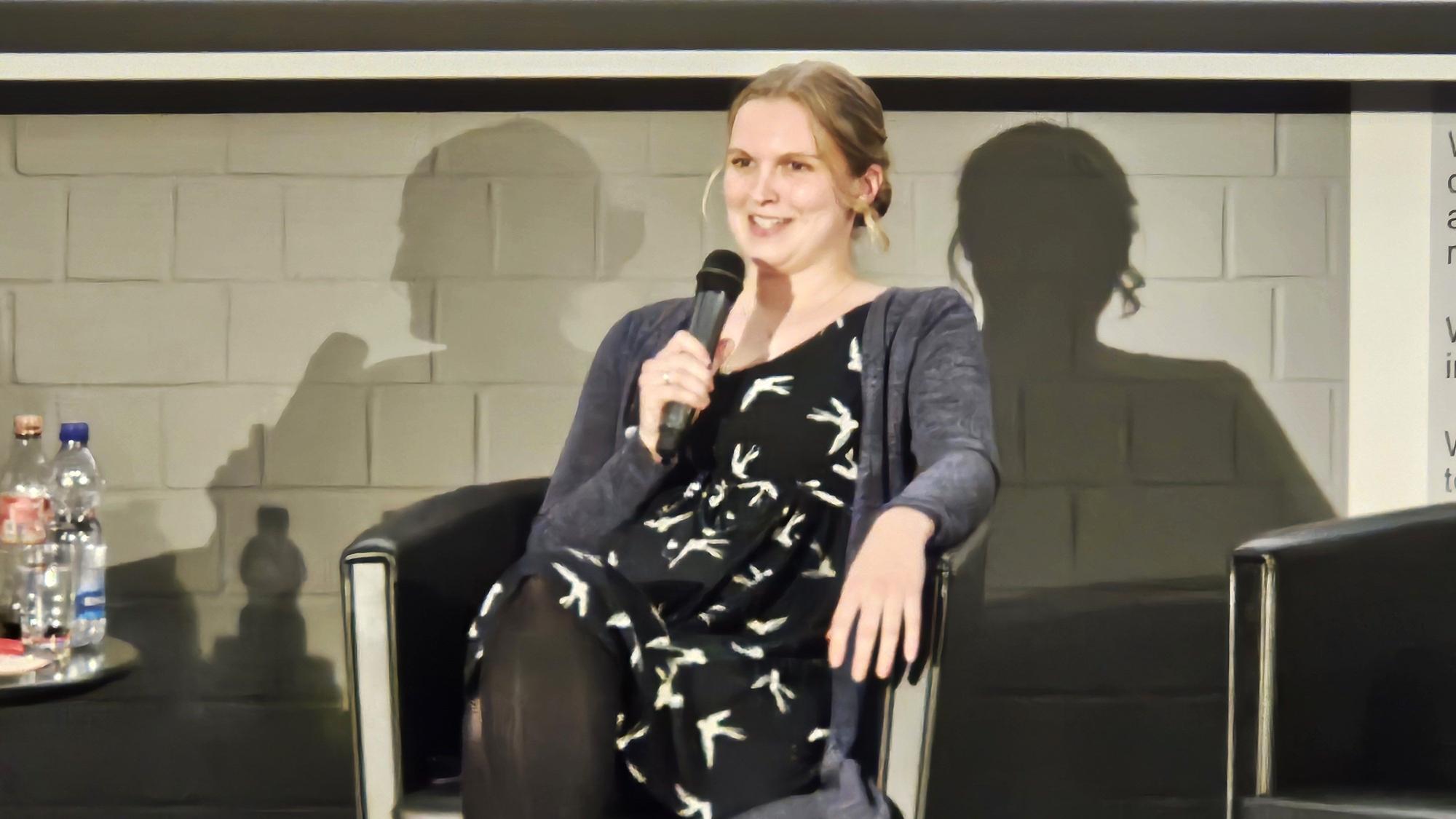 Maria Schmidt sitzt mit einem Mikro in der Hand auf der Bühne.