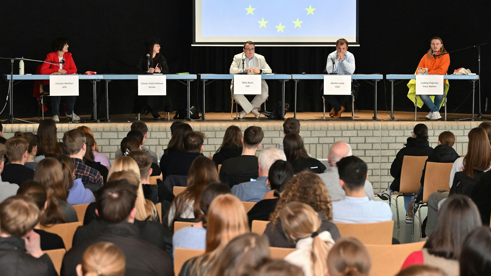 Jugendliche sitzen im Publikum, auf der Bühne sitzen Politiker mit Namensschildern. Hinter ihnen die Europafahne.