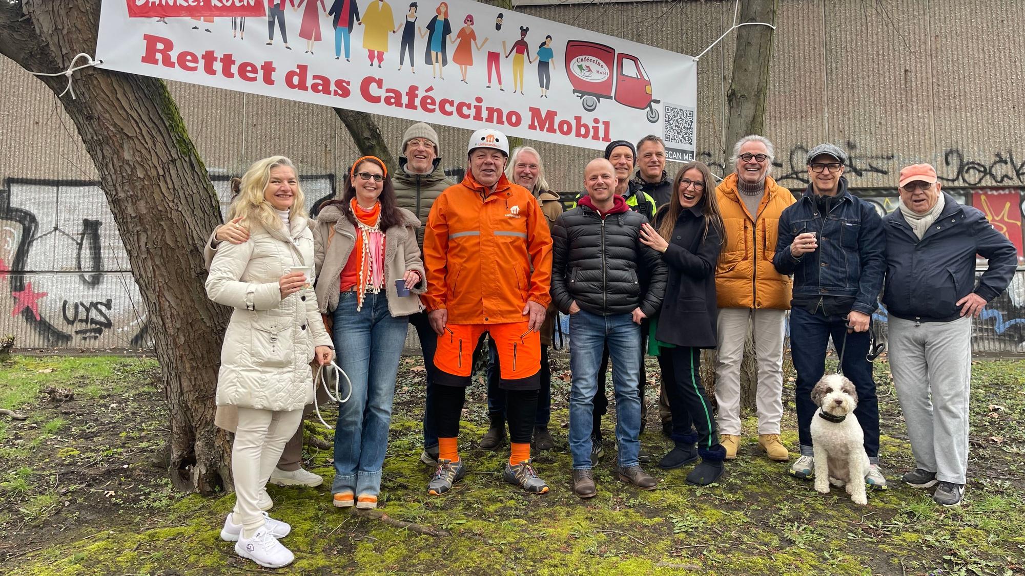 Viele Menschen stehen unter Bäumen. Zwischen zwei Stämmen haben sie ein Banner gespannt, auf dem steht: Rettet das Cafeccino-Mobil.
