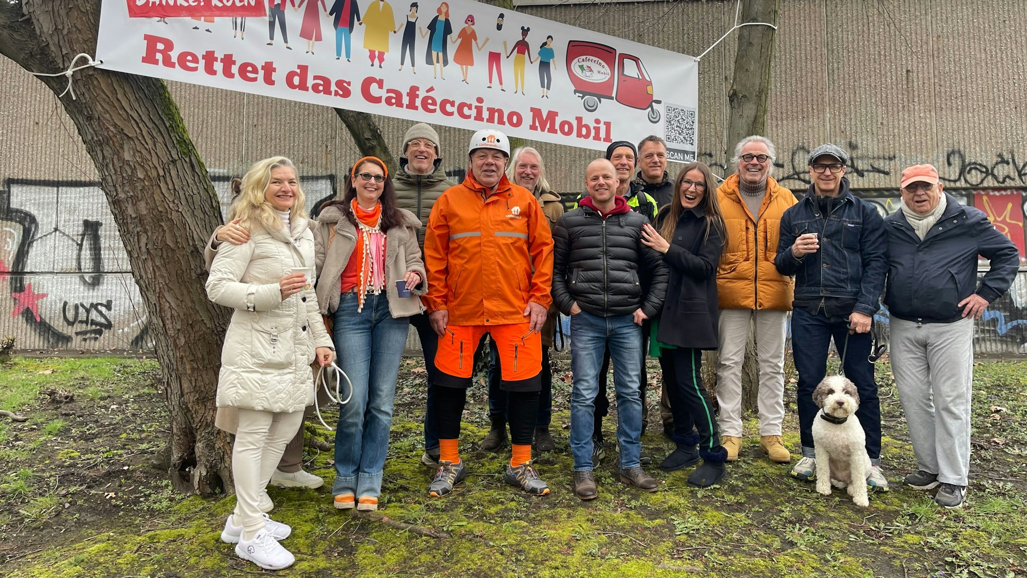 Viele Menschen stehen unter Bäumen. Zwischen zwei Stämmen haben sie ein Banner gespannt, auf dem steht: Rettet das Cafeccino-Mobil.