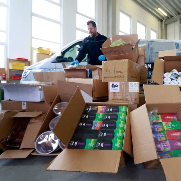 Kistenweise sichergestellte Tabakprodukte liegen in einer Lagerhalle, dahinter stehen zwei Fahrzeuge und ein Beamter des Zollamts.