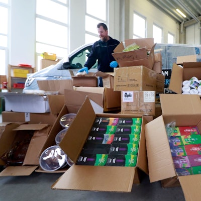 Kistenweise sichergestellte Tabakprodukte liegen in einer Lagerhalle, dahinter stehen zwei Fahrzeuge und ein Beamter des Zollamts.