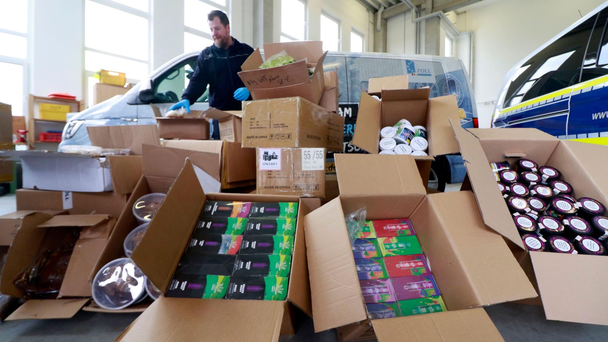 Kistenweise sichergestellte Tabakprodukte liegen in einer Lagerhalle, dahinter stehen zwei Fahrzeuge und ein Beamter des Zollamts.