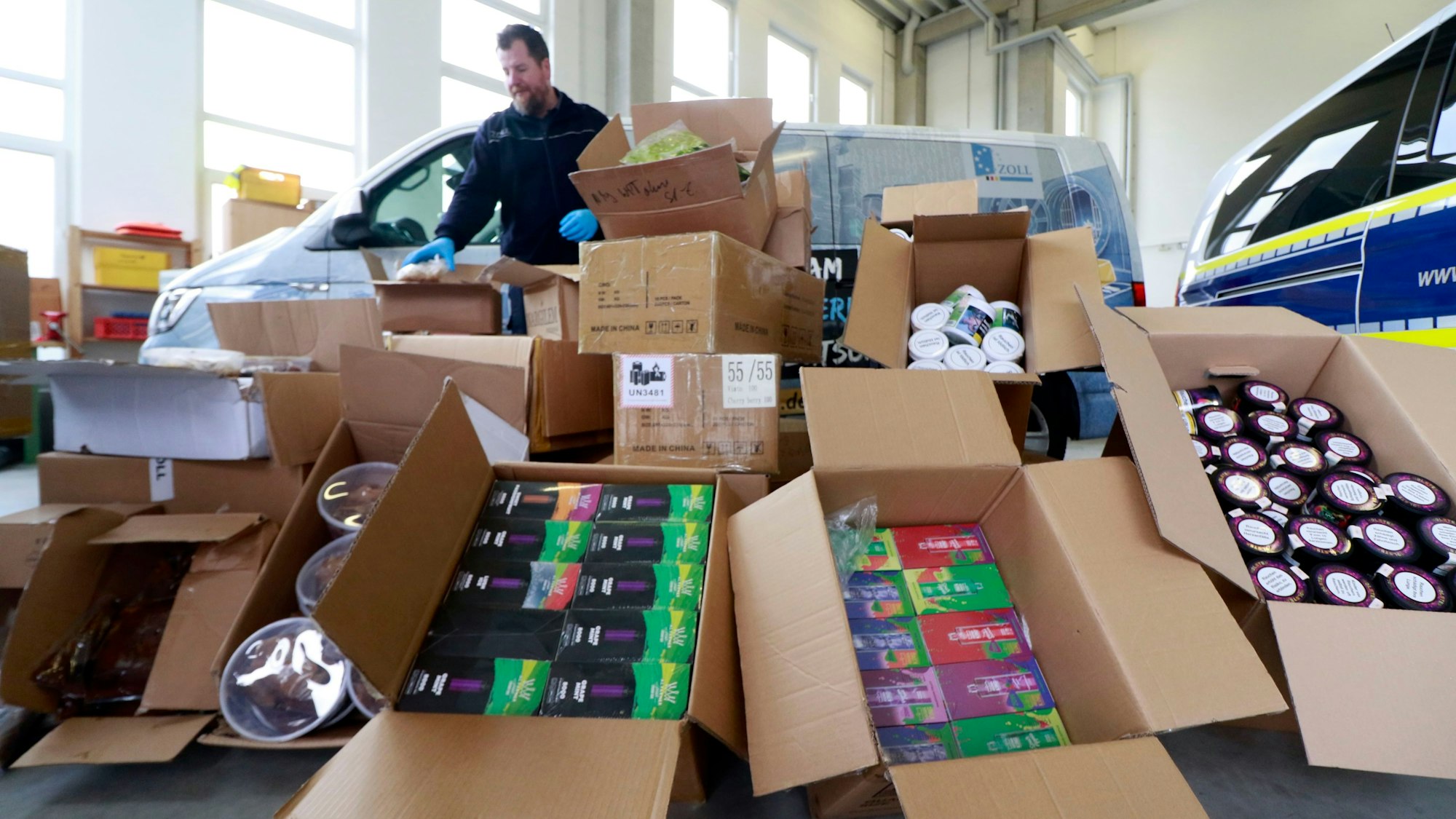 Kistenweise sichergestellte Tabakprodukte liegen in einer Lagerhalle, dahinter stehen zwei Fahrzeuge und ein Beamter des Zollamts.