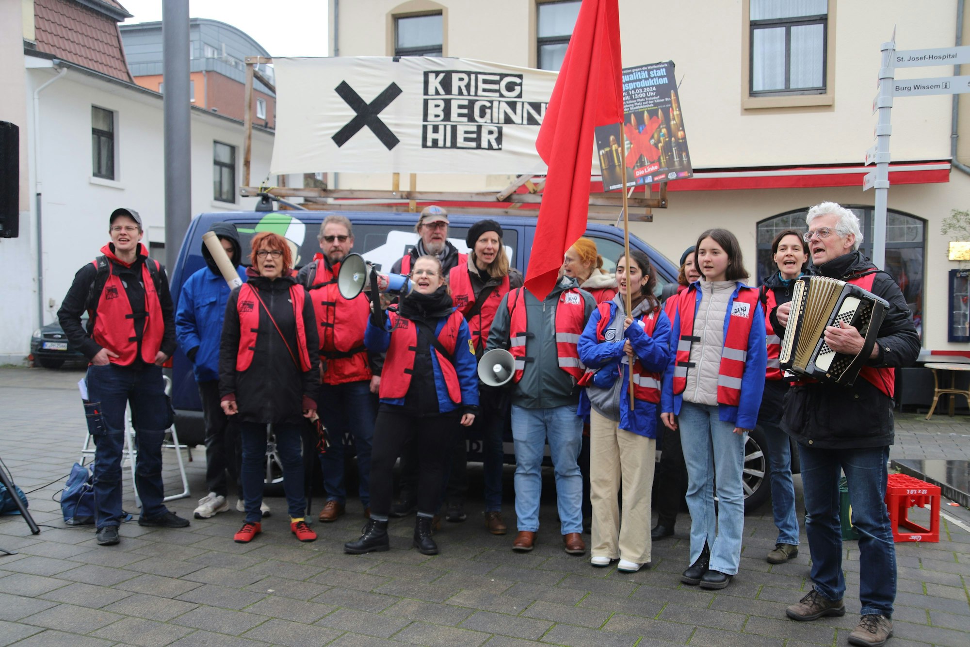 „Krieg beginnt hier“; heißt es auf einem Schild an einem Fahrzeug. Davor steht eine Gruppe von Menschen.