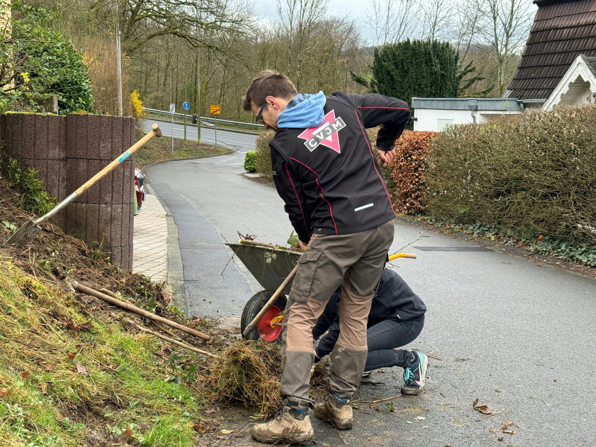 Zwei junge Menschen in Arbeitskleidung säubern eine Bäuschung neben einer Straße.