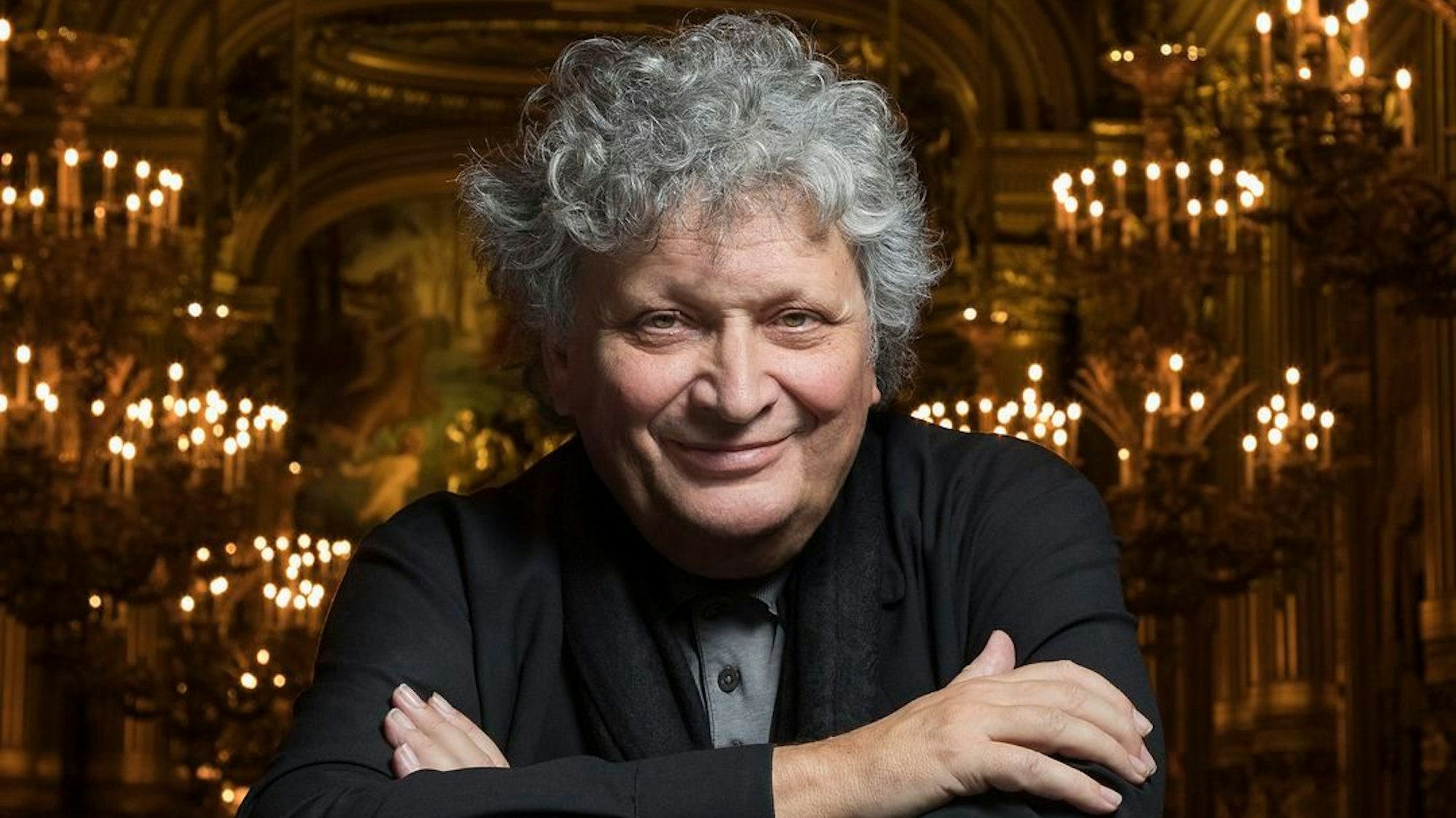 Auf dem Bild ist der Dirigent zu sehen, er hält die arme überkreuz. Er hat weißes, lockiges Haar und trägt einen schwarzen Anzug. Im Hintergrund ein sakral anmutendes Gebäude mit vielen Kerzen.