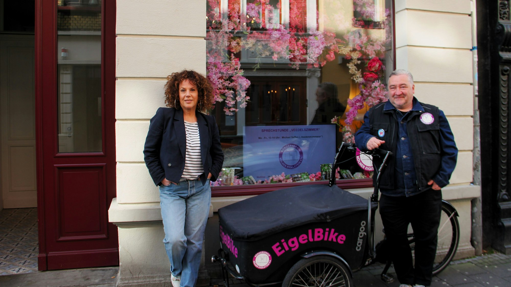 Eine Frau mit gelocktem Haar und ein Mann mit grauem Haar stehen neben einem Lastenrad mit der Aufschrift EigelBike.
