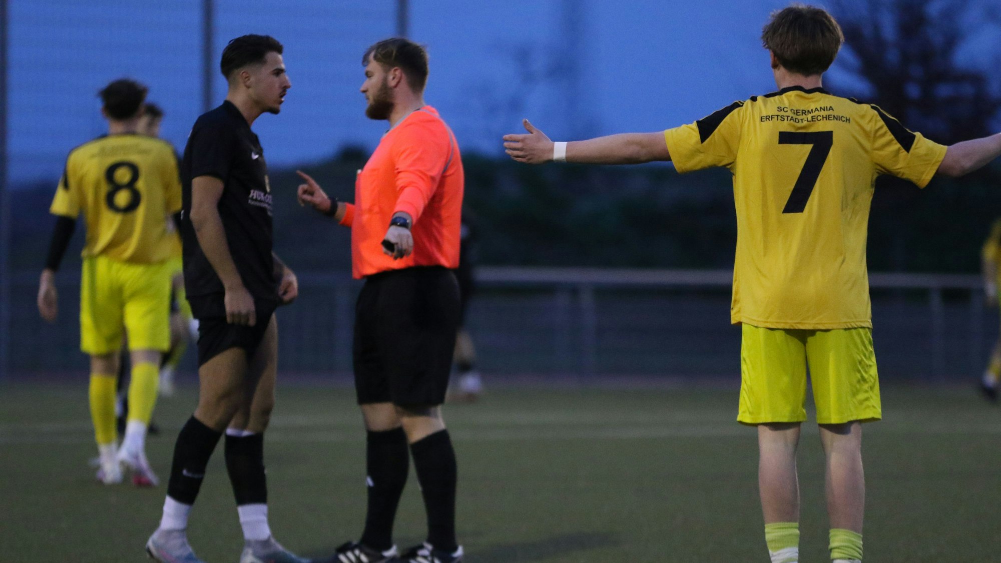 Schiedsrichter Eric Strohmeier spricht auf dem Fußballplatz mit dem JSG-Spieler Arber Jashanica, ein Erftstädter Akteur beobachtet die Szene mit ausgebreiteten Armen.
