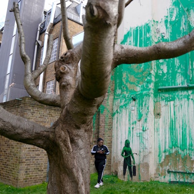 Eine Frau im Trainingsanzug steht auf einem Garten neben einer Wand, die mit Kunst besprüht wurde.