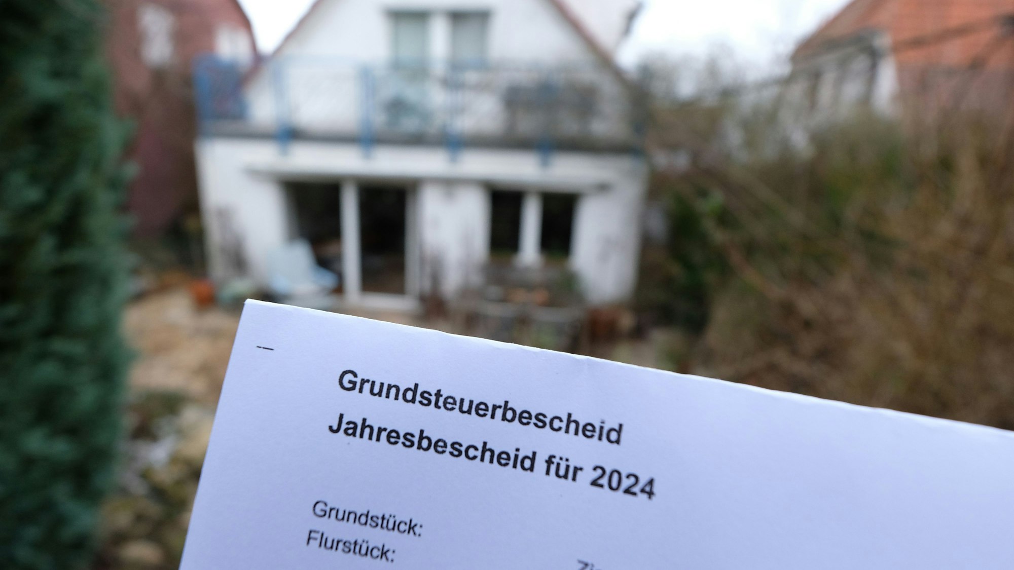 Ein Grundsteuerbescheid für 2024 wird vor einem Haus mit Garten hochgehalten.