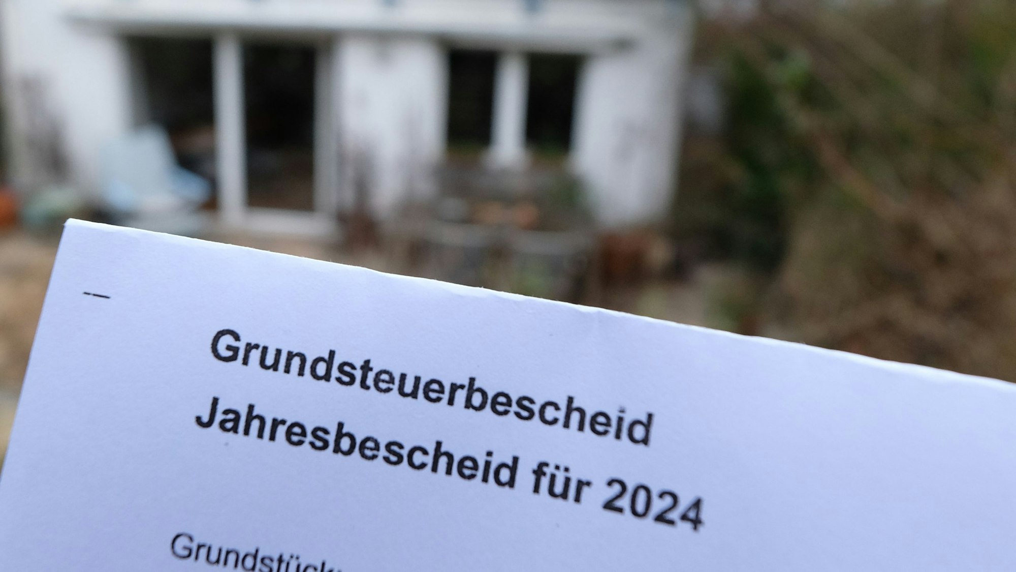Ein Grundsteuerbescheid für 2024 wird vor einem Privatgrundstück hochgehalten. Für die neue Berechnung der Grundsteuer müssen bundesweit fast 36 Millionen Grundstücke neu bewertet werden.