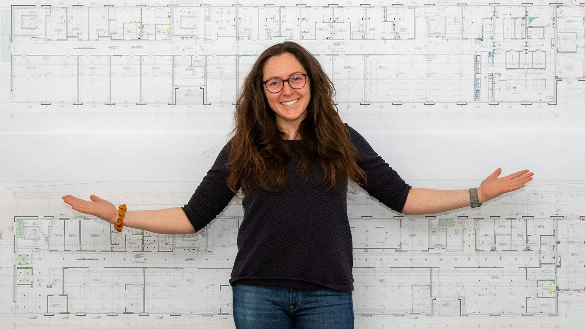 Christina Pötz breitet die Arme aus und steht vor den Bauplänen des neuen Gebäudes.