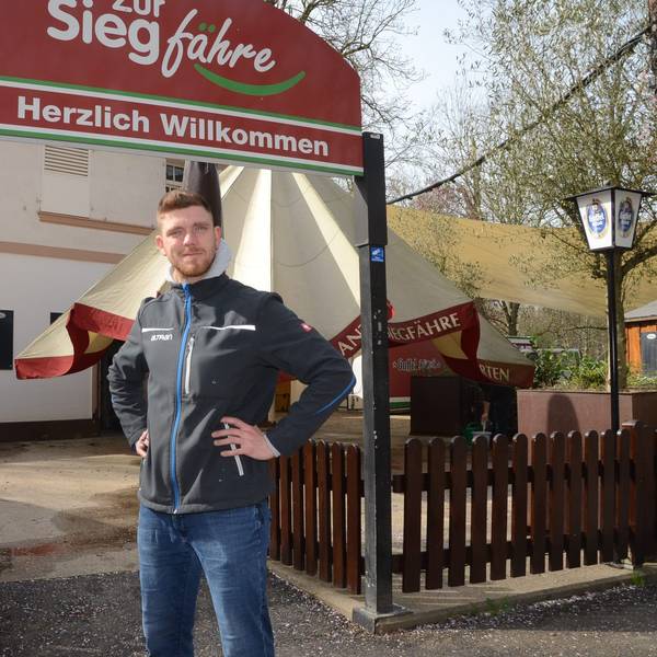 Ein Mann steht vor dem Eingang zu einem Biergarten. „Zur Siegfähre“ steht auf dem Schild, „Herzlich willkommen“ in der unteren Zeile.