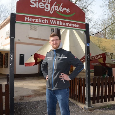 Ein Mann steht vor dem Eingang zu einem Biergarten. „Zur Siegfähre“ steht auf dem Schild, „Herzlich willkommen“ in der unteren Zeile.