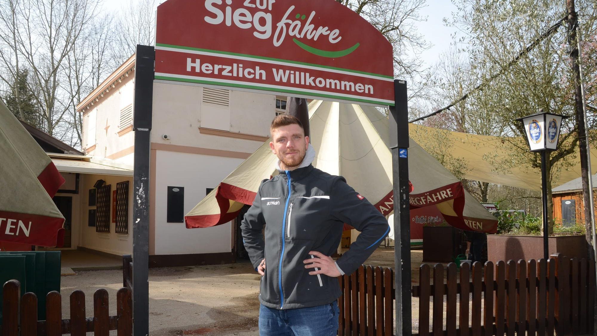 Ein Mann steht vor dem Eingang zu einem Biergarten. „Zur Siegfähre“ steht auf dem Schild, „Herzlich willkommen“ in der unteren Zeile.
