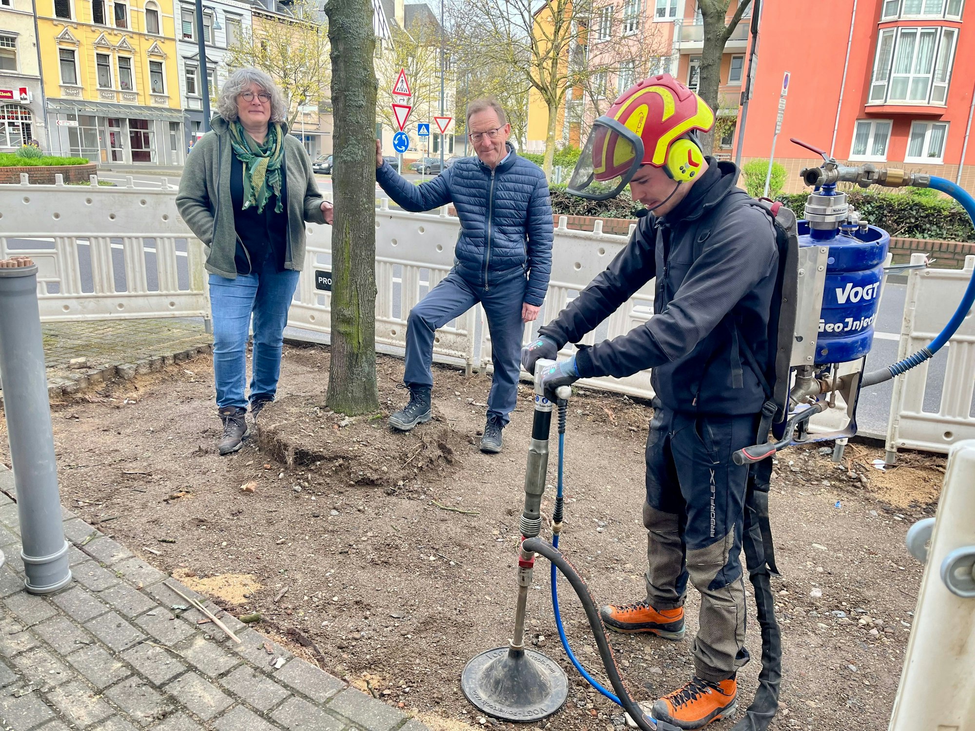 Ein Mann lockert Boden an einem Straßenbaum auf. Eine Frau und ein Mann schauen zu.