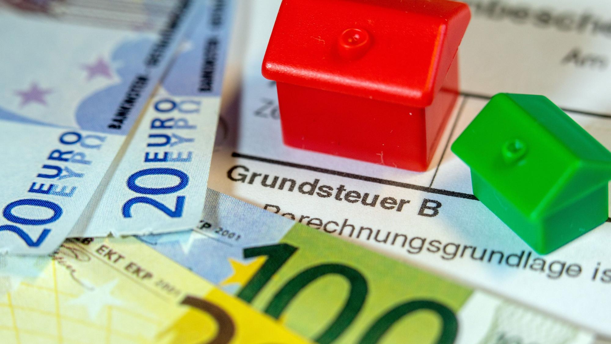Ein Spielzeughaus und die Nachbildung von Euro-Geldscheinen liegen auf einem Abgabenbescheid für die Entrichtung der Grundsteuer