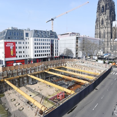 Das Bild zeigt eine Baustellengrube vor einigen Gebäuden, im Hintergrund ist der Dom zu sehen.