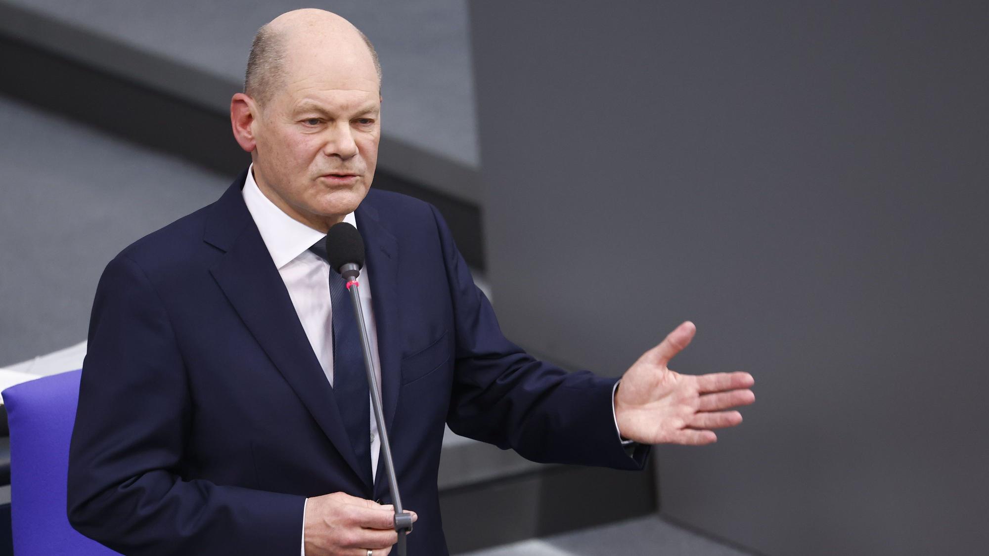 Olaf Scholz während seiner Bundestagsrede am 13. März, in der er die Nichtentsendung von Taurus-Marschflugkörpern verteidigte. Er steht vor einem Mikrofon, den linken Arm und die geöffnete linke Hand in einer erklärenden Geste von sich gestreckt. Sein Gesichtsausdruck ist ruhig und gefasst.