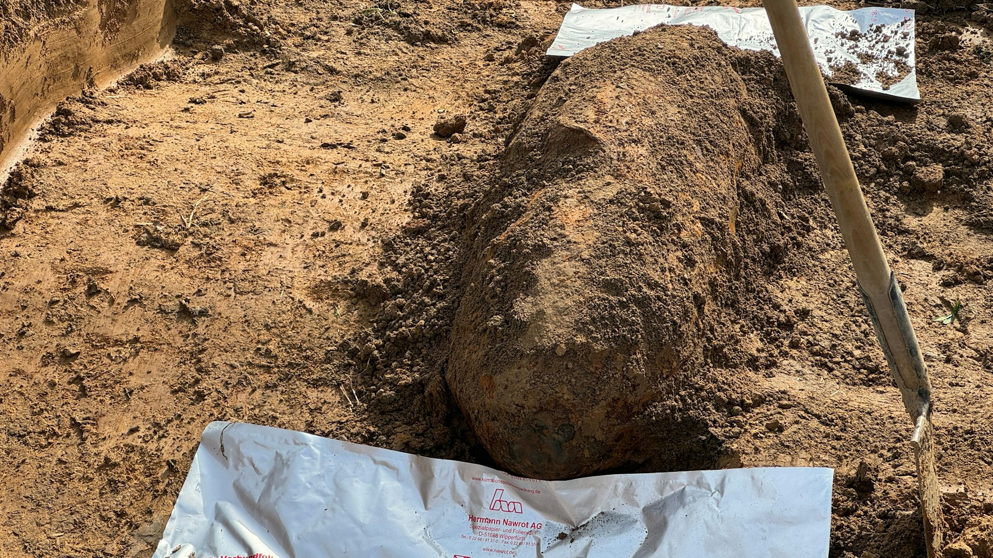 Ein mit Erde bedeckter Blindgänger liegt in einem Graben, daneben steht eine Schaufel.