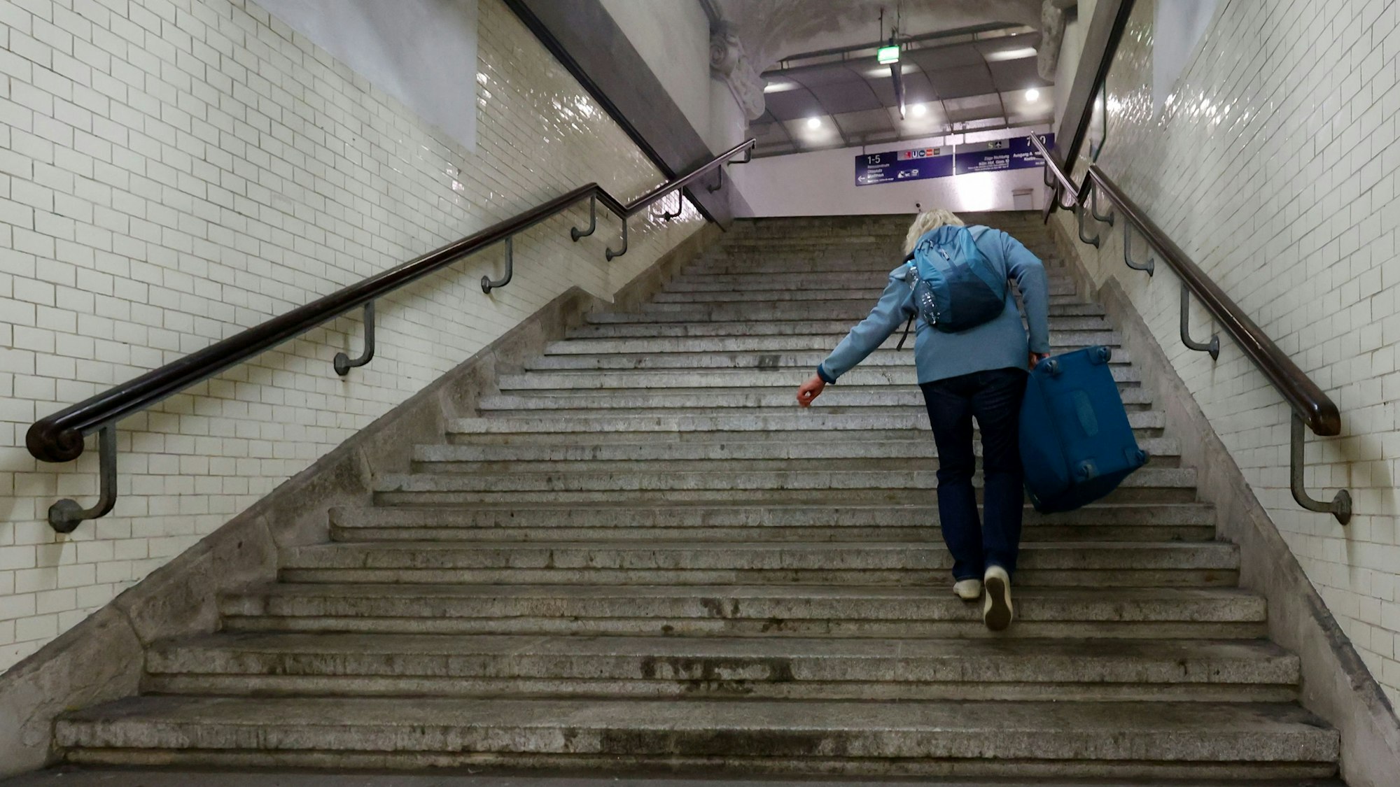 Im Bahnhof Köln-Deutz führt eine steile Treppe zu den S-Bahn-Gleisen. Eine Reisende mit Rucksack steigt mit Koffer die Treppe hinauf.