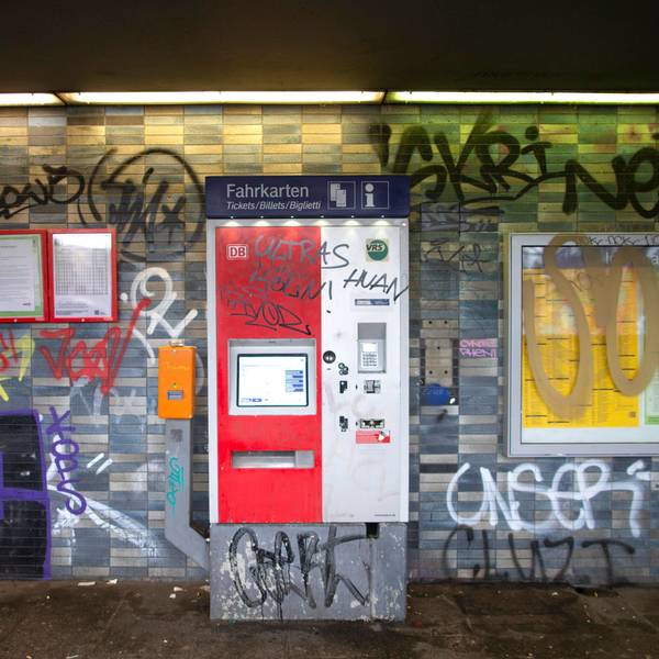 Eine gekachelte Wand in einem Bahnhof sowie ein Fahrkartenautomat und Fahrplanaushänge sind mit Graffiti und Schriftzügen beschmiert.