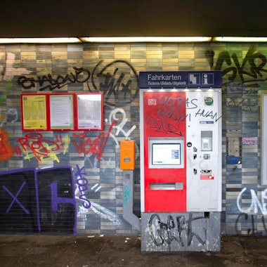 Eine gekachelte Wand in einem Bahnhof sowie ein Fahrkartenautomat und Fahrplanaushänge sind mit Graffiti und Schriftzügen beschmiert.