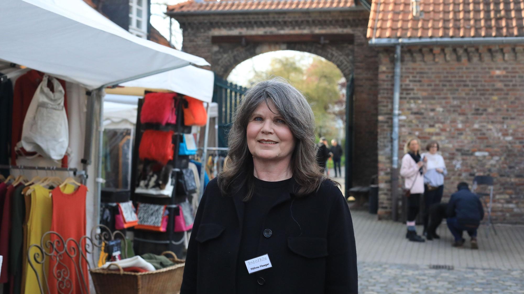 Eine Frau steht im Hof eines großen Gutshofs, links neben ihr ist ein Verkaufsstand zu sehen.