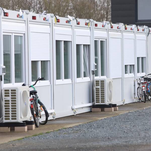 Wohncontainer auf einem Schotterplatz. Vor den Containern stehen Lüftungsgeräte, an einige sind Fahrräder angelehnt.