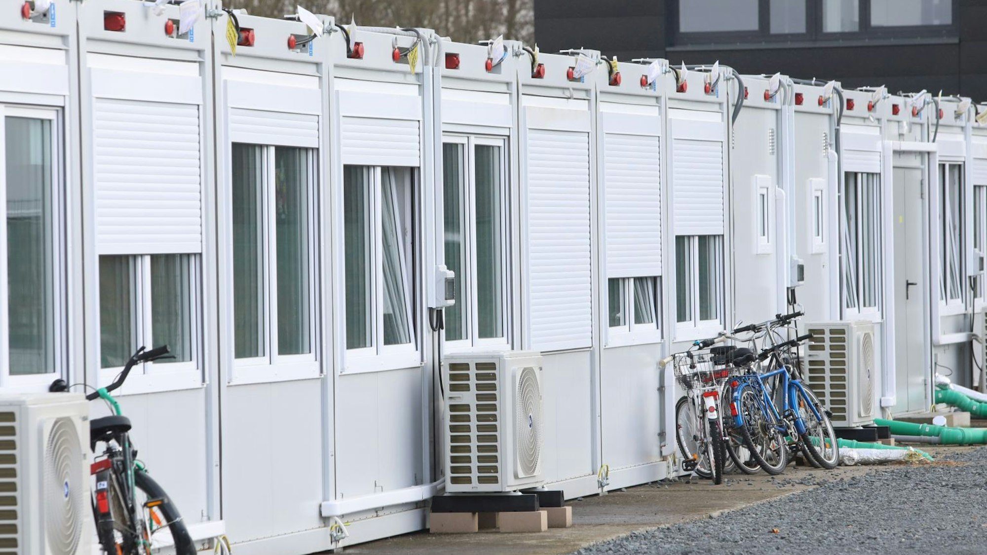 Wohncontainer auf einem Schotterplatz. Vor den Containern stehen Lüftungsgeräte, an einige sind Fahrräder angelehnt.