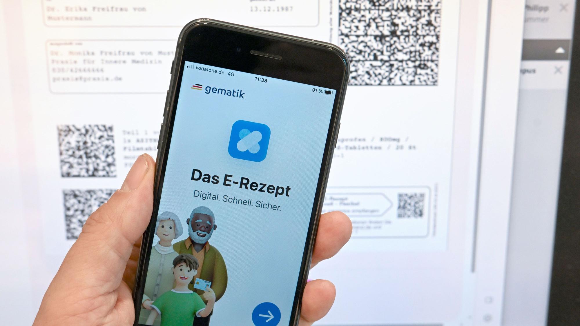 Eine App, die das E-Rezept anzeigen kann, wird in einer Apotheke vor einem Bildschirm gehalten, der Hinweise zu den E-Rezepten anzeigt.