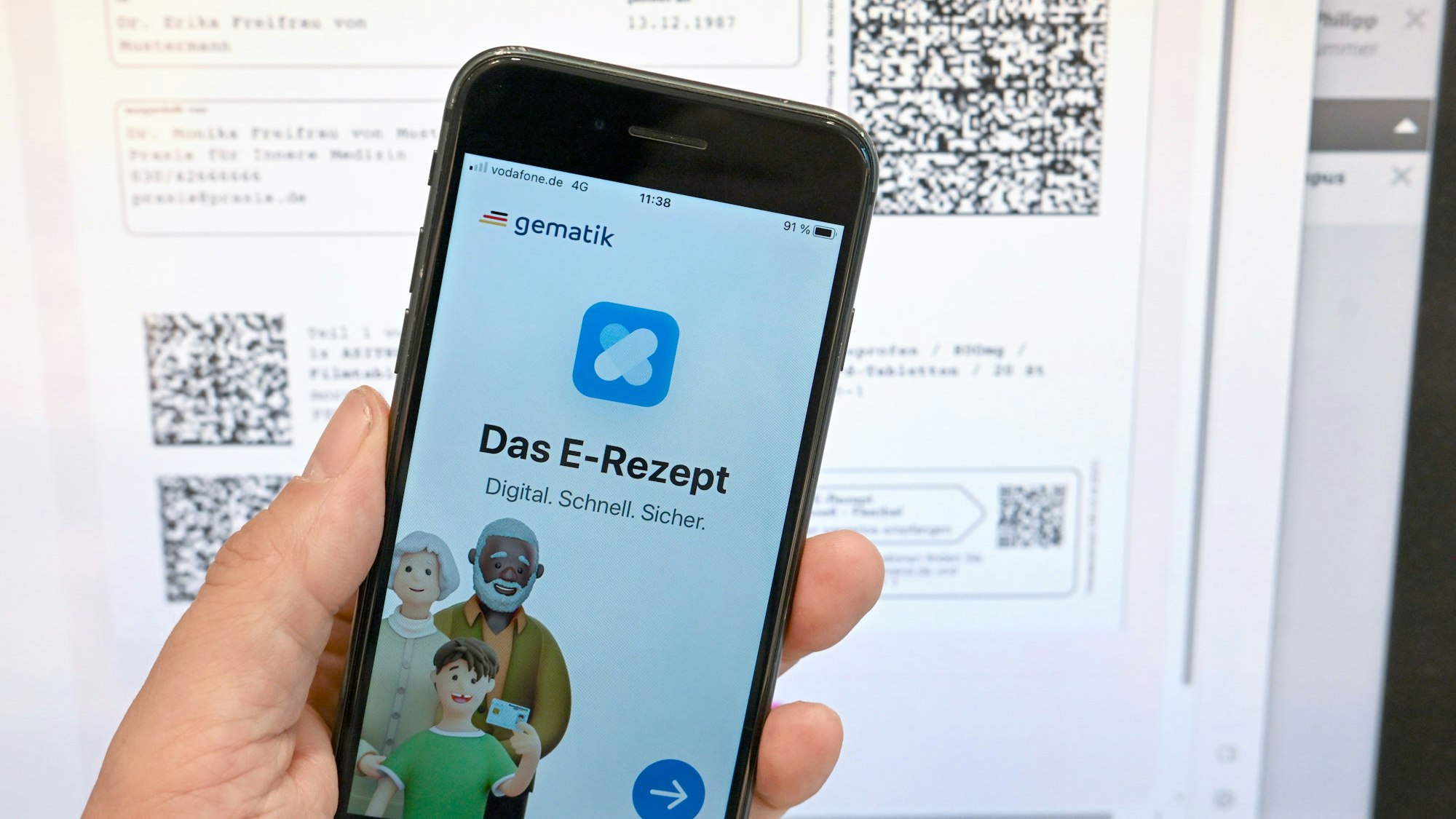 Eine App, die das E-Rezept anzeigen kann, wird in einer Apotheke vor einem Bildschirm gehalten, der Hinweise zu den E-Rezepten anzeigt.