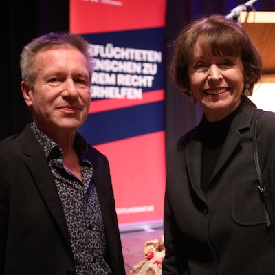Ein Mann und eine Frau vor einem Plakat mit der Aufschrift „Geflüchteten Menschen zu ihrem Recht verhelfen“