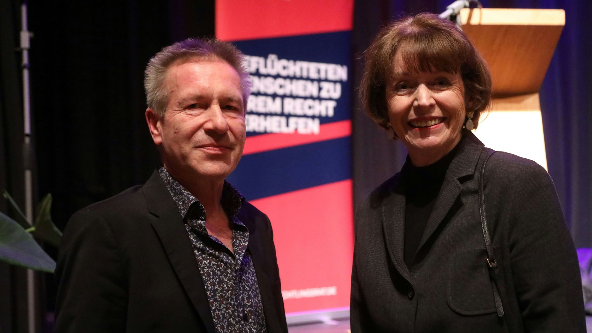 Ein Mann und eine Frau vor einem Plakat mit der Aufschrift „Geflüchteten Menschen zu ihrem Recht verhelfen“