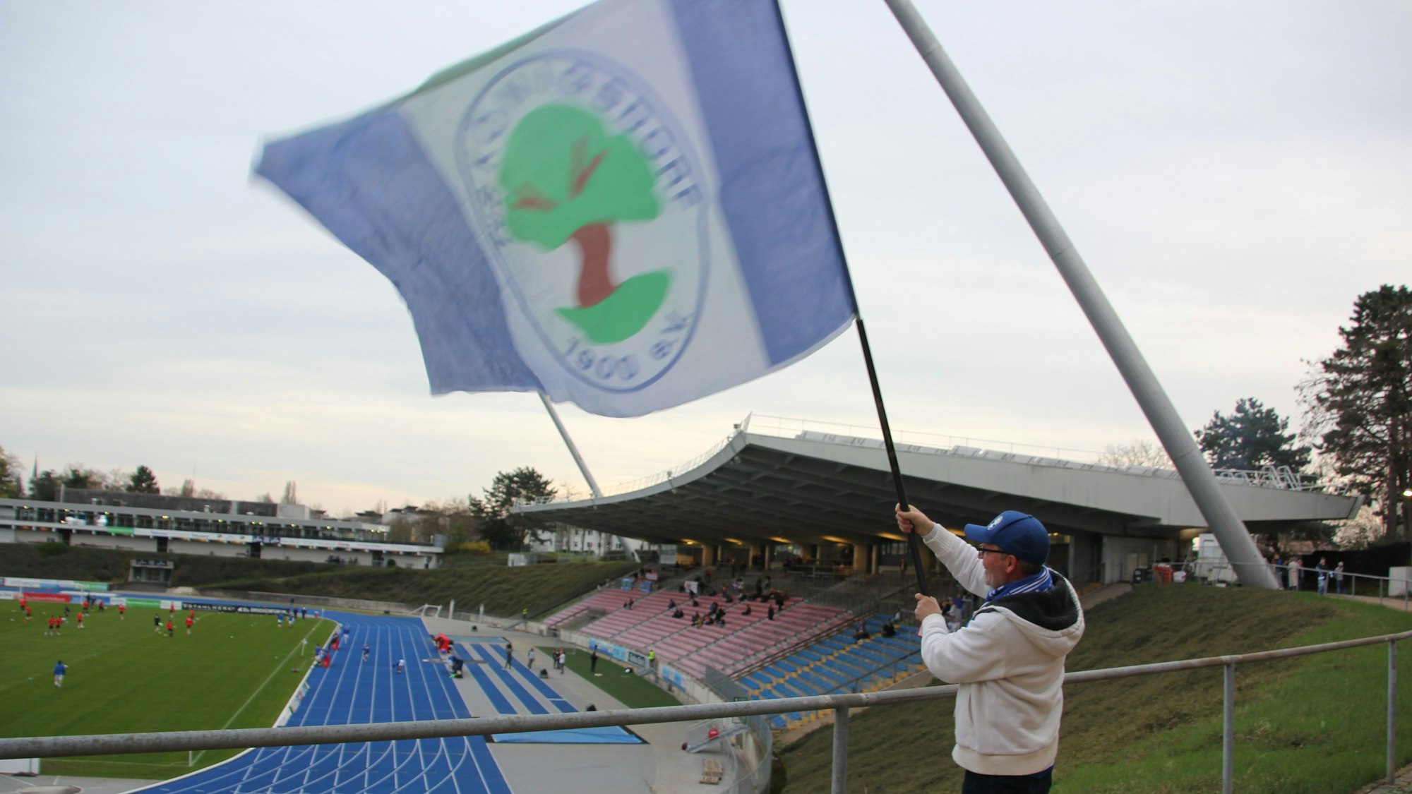 Ein Fan schwenkt eine große Fahne des TuS Blau-Weiß Königsdorf. Im Hintergrund ist die Haupttribüne des Bonner Stadions zu sehen.