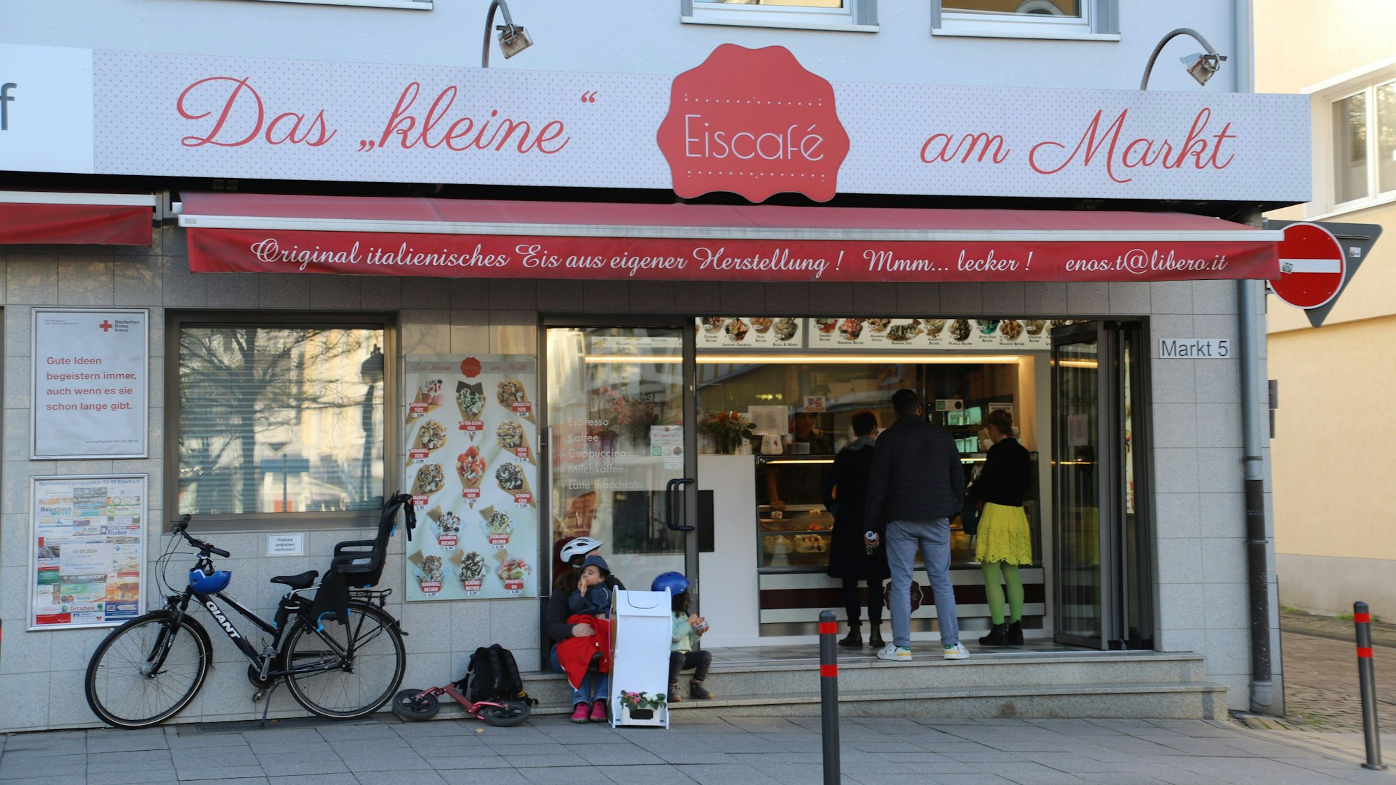 Das kleine Eiscafe am Markt in Eitorf in der Außenansicht. Menschen bestellen an der Eistheke, links im Bild ist ein Fahrrad mit Kindersitz an die Hauswand gelehnt.