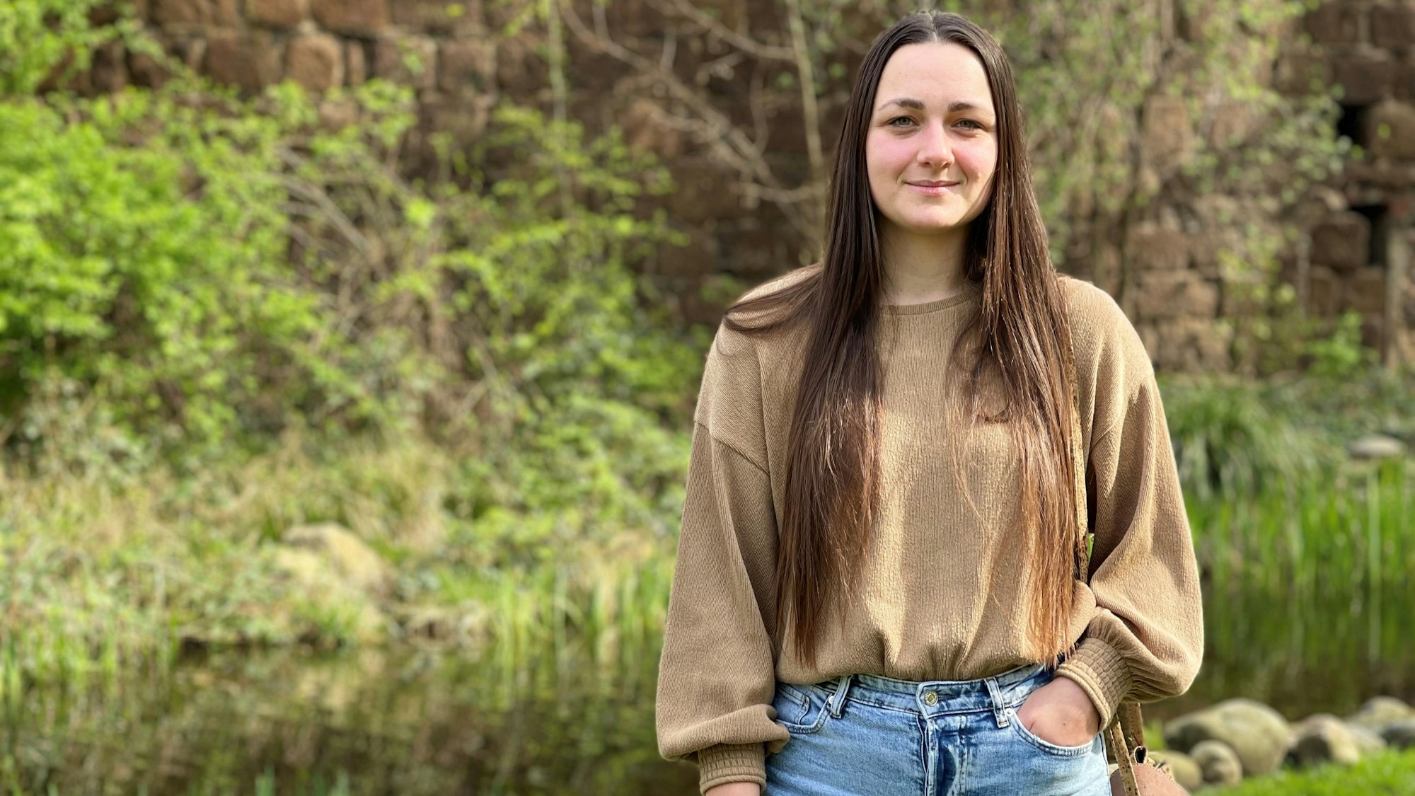 Eine junge Frau mit langen, dunklen Haaren steht vor grünen Pflanzen.