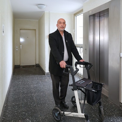 Zu sehen ist Halis Yildirim aus Bocklemünd in der Vorderansicht vor einem Aufzug mit Rollator stehend.